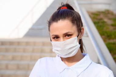 Beyaz tişörtlü beyaz esmer bir kızın şehrin güneşli bir gününde binanın arka planında çekilmiş bir fotoğrafı. Tıbbi koruyucu maskeli bir model..
