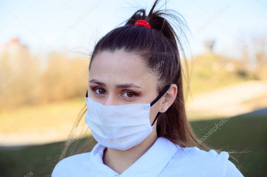Photo Caucasian young brunette girl in a mask from a virus in a white T-shirt outdoors in the afternoon in the city on a street background.
