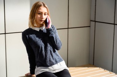 Şehrin güneşli bir gününde, gri kazaklı beyaz bir kızın bir binanın arka planında çekilmiş fotoğrafı. Model akıllı telefon kullanıyor..