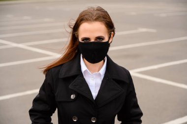 Bir otoparkın arka planında açık havada siyah bir ceketle yürürken şehir sokaklarında siyah maskeli esmer bir kızın portresi.