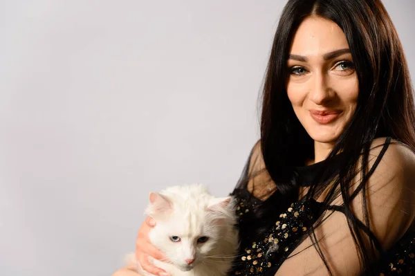 Fashion model glamorous caucasian brunette girl posing with a white cat in her arms on a white background.