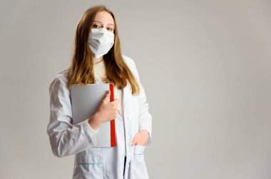 Koruyucu maske ve beyaz önlüklü güzel sarışın bir doktorun stüdyo portresi. Bir kadın elinde bir klasörle beyaz bir arka planda duruyor..