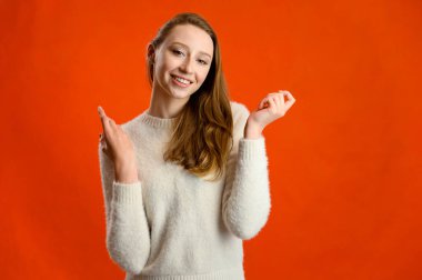 Model konuşuyor, mutlu duygular gösteriyor. Stüdyo fotoğrafı beyaz kazaklı beyaz bir kızın kırmızı arka planda kameranın önünde duruşu..