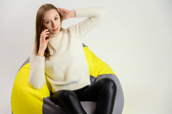 Studio portrait of a happy caucasian girl in a white sweater sitting on a white background talking with a phone in her hands