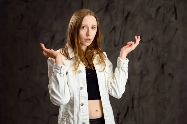 Model is talking, showing emotions. Studio photo of a happy caucasian girl in a white jacket stands in front of the camera on a gray background.