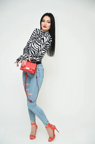 Model glamorous girl in blue jeans and a striped blouse stands right with a red bag on a white background. Vertical photo of a fashionable caucasian brunette.