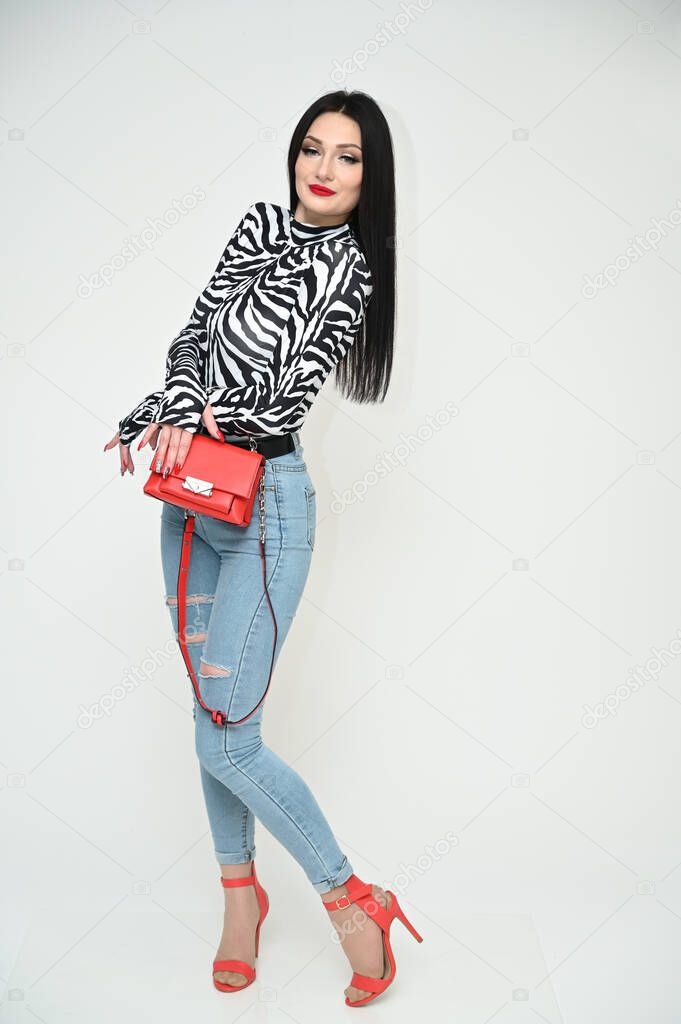 Model glamorous girl in blue jeans and a striped blouse stands right with a red bag on a white background. Vertical photo of a fashionable caucasian brunette.