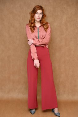 Manken pembe gömlek, kırmızı pantolon ve gri ayakkabı giyiyor. Tam boy dikey portre düz beyaz, kızıl saçlı, bej arka planda güzel bir kadın..