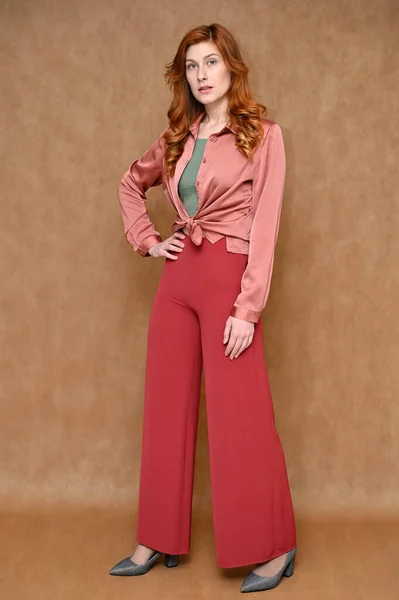 The model is dressed in a pink shirt and red pants, gray shoes. Full length vertical portrait straight Caucasian standing red hair pretty young woman on a beige background.