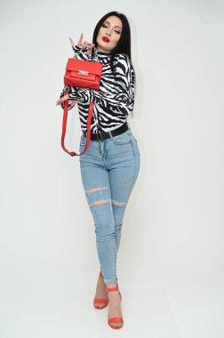 Mükemmel makyajlı beyaz tenli bir esmerin tam boy yatay portresi. Çizgili bluzlu ve kot pantolonlu göz kamaştırıcı kız stüdyoda beyaz bir arka planda kırmızı bir çanta gösteriyor..