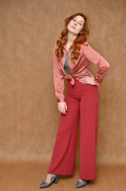 Tam boy dikey portre, bej arka planda beyaz, kızıl saçlı hoş bir kadının duruşu. Manken pembe gömlek, kırmızı pantolon ve gri ayakkabı giyiyor..