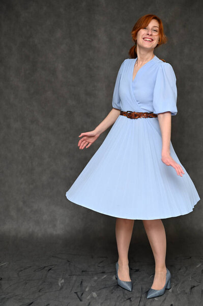 Девушка-модель позирует в студии на сером фоне. Полнометражный портрет рыжеволосой девушки в синем платье
.
