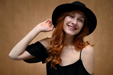 Bej arka planda siyah şapkalı, uzun kızıl saçlı mutlu bir kadının portresi. Gülümseyen bir model.