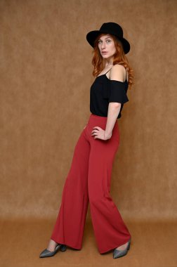 Stüdyoda bej bir arka planda elinde şapkayla poz veren kız mankeni. Kızıl saçlı, siyah bluzlu, kırmızı pantolonlu güzel bir kadının tam boy portresi..