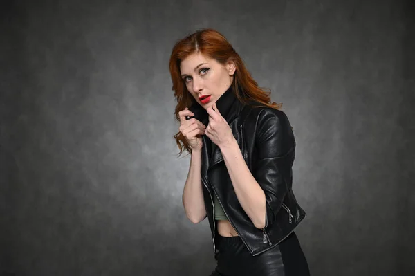 Pretty Model with great makeup posing on a gray background in the studio. Horizontal portrait of a red-haired young woman in a black jacket and green t-shirt.