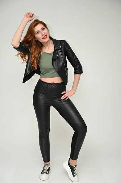 Vertical portrait on a white background of a pretty red-haired young woman. A full-length model with excellent makeup poses in a black jacket and trousers in the studio.
