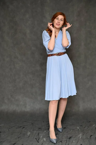 The model demonstrates standing in full growth different poses. Vertical portrait of a young pretty red-haired woman in a blue dress on a gray background.