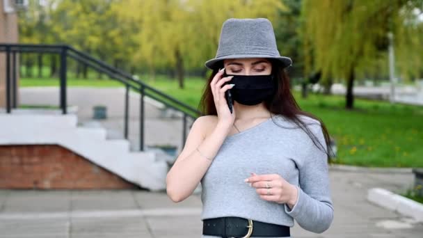 街の公園の自然を背景にした医療用マスクで電話で話しているかなり笑顔ブルネットの女の子のビデオポートレート — ストック動画
