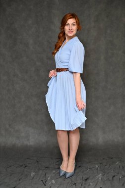 Model farklı pozlar veriyor. Gri arka planda mavi elbiseli, güzel, kızıl saçlı bir kadının dikey portresi..