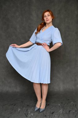 Model farklı pozlar veriyor. Gri arka planda mavi elbiseli, güzel, kızıl saçlı bir kadının dikey portresi..
