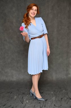 Model, elinde bir çiçekle farklı pozlarda durduğunu gösteriyor. Gri arka planda mavi elbiseli güzel, kızıl saçlı bir kadının dikey portresi..