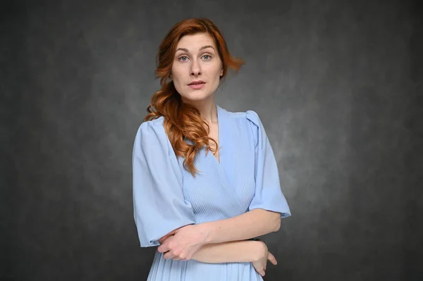 Egy fiatal, csinos, vörös hajú nő portréja, kék ruhában, szürke háttérrel. A modell különböző érzelmeket mutat.. — Stock Fotó