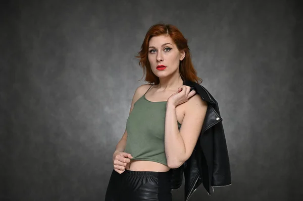 Το μοντέλο δείχνει διαφορετικά συναισθήματα αλλάζοντας πόζες. Πορτρέτο μιας νεαρής όμορφης κοκκινομάλλας με πράσινο μπλουζάκι και μαύρο μπουφάν σε γκρι φόντο. — Φωτογραφία Αρχείου