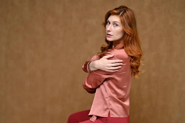 Ein hübsches Model mit langen Haaren trägt eine rote Hose und eine rosafarbene Bluse. Porträt einer jungen rothaarigen Frau aus dem Kaukasus, die auf einem Ständer auf beigem Hintergrund in verschiedenen Posen sitzt. — Stockfoto