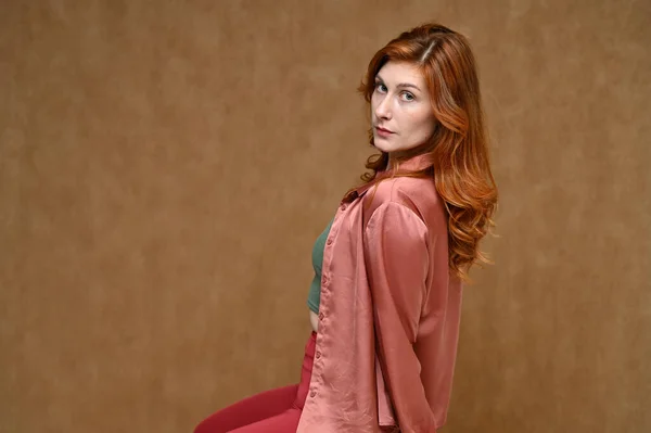 Egy fiatal kaukázusi vörös hajú nő portréja, bézs háttérrel, különböző pózokban. Egy szép modell hosszú hajjal piros nadrágban és rózsaszín blúzban.. — Stock Fotó