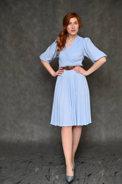 Gri arka planda mavi elbiseli güzel, kızıl saçlı bir kadının tam boy dikey portresi. Model farklı pozlar gösterir.