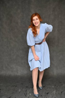 Gri arka planda mavi elbiseli güzel, kızıl saçlı bir kadının tam boy dikey portresi. Model farklı pozlar gösterir.