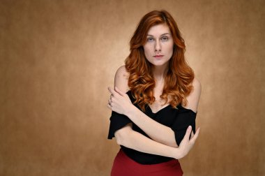 Siyah bluzlu, bej arka planda uzun kızıl saçlı beyaz bir kadının stüdyo fotoğrafı. Model farklı duygular gösteriyor..