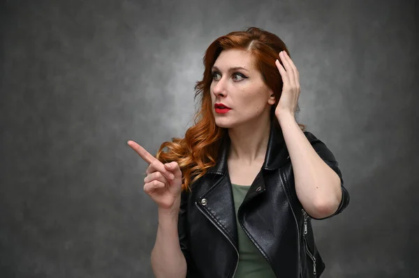 Pretty model actress posing with different emotions on a gray background in the studio. Studio photo of a young caucasian woman with long red hair in a black jacket.
