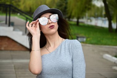 Gri elbiseli, elinde gözlüklerle güzel, esmer bir kızın fotoğrafı. Şehir parkında poz veren bir model.
