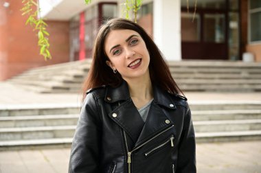 Çeşitli pozlarda siyah ceketli, beyaz, tatlı, esmer bir kızın portresi. Şehirde poz veren bir model. Baharda açık havada, gülümseyerek..