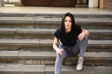Bahar havasında açık havada tam boy portre, kot pantolon ve siyah tişörtlü beyaz tenli kız. Modaya uygun bir model binanın merdivenlerinde oturuyor..
