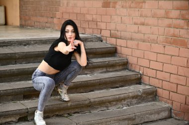 Bahar havasında açık havada tam boy portre, kot pantolon ve siyah tişörtlü beyaz tenli kız. Binanın arka planında poz veren model..