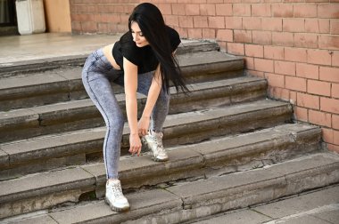 Bahar havasında açık havada tam boy portre, kot pantolon ve siyah tişörtlü beyaz tenli kız. Binanın arka planında poz veren model..