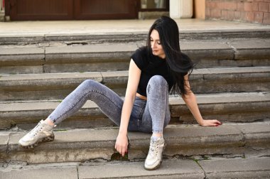 Bir model bir binanın merdivenlerinde oturur. Bahar havasında tam boy portre, beyaz tenli, kot pantolon ve siyah tişörtlü kız..