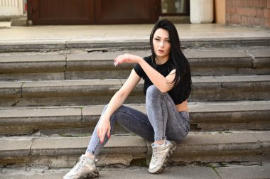 Bahar havasında açık havada tam boy portre, kot pantolon ve siyah tişörtlü beyaz tenli kız. Binanın merdivenlerinde bir model oturuyor..