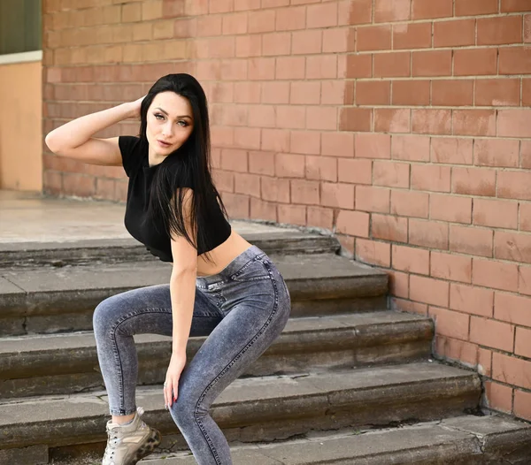 Model posing on the background of the stairs of the building. Full-length portrait in spring weather outdoors, caucasian brunette girl with a slim figure in jeans and a black T-shirt.