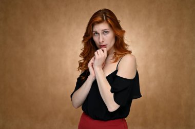Model aktris stüdyoda konuşurken farklı duygular gösterir. Siyah bluzlu, uzun kızıl saçlı, bej arkaplanlı, asgari makyajlı beyaz bir kadının fotoğrafı..