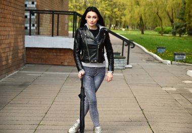 Bir model elinde şemsiyeyle şehir parkına karşı poz veriyor. Bahar havasında tam boy portre, siyah ceket ve kot pantolon giyen Kafkasyalı modaya uygun esmer kız..
