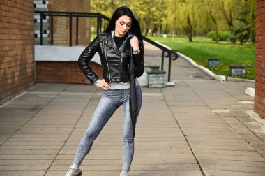 Baharda açık havada tam boy portre, siyah ceketli ve kot pantolonlu esmer kız. Bir model elinde şemsiyeyle şehir parkına karşı poz veriyor..