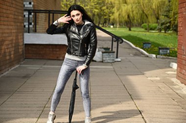 Şehir parkının arka planında şemsiyeyle poz veren bir model. Siyah ceketli ve kotlu, açık havada duran beyaz, modaya uygun esmer bir kızın tam boy portresi..