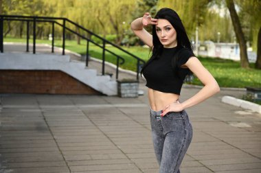 Fitness modeli, güzel havada bir şehir parkının arka planında duruyor. Bahar havasında, beyaz tenli, zayıf vücutlu esmer bir kız zevkle poz veriyor..