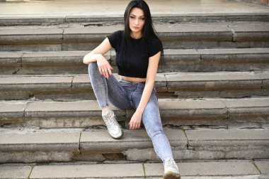 Bahar havasında açık havada tam boy portre, kot pantolon ve siyah tişörtlü beyaz tenli kız. Modaya uygun bir model binanın merdivenlerinde oturuyor..