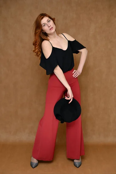 ベージュ地に全長の肖像写真黒のブラウス 赤いズボンを着た白人のかなり若い女性 モデルポーズで彼女の手に帽子とともに長い美しい赤い髪 — ストック写真