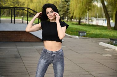 Kot pantolon ve siyah tişört giymiş ince vücutlu açık hava esmer kız portresi. Model, güzel havada bir şehir parkının arka planına karşı duruyor..