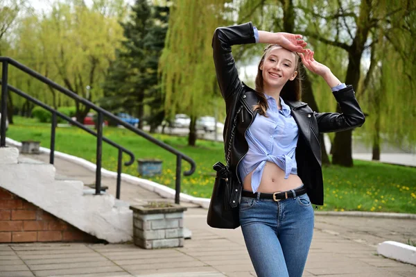 模特儿站在城外的春天公园里 一个穿着黑色夹克和牛仔裤笑着的年轻漂亮姑娘的照片 — 图库照片
